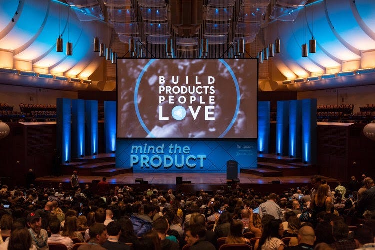 Foto mostra um auditório cheio de pessoas sentadas, um palco vazio e um telão, em que está escrito: "Build Products People Love. Mind The Product" ("Construindo produtos que as pessoas amam. Mind The Product"). Foto mostra um auditório cheio de pessoas sentadas, um palco vazio e um telão, em que está escrito: "Build Products People Love. Mind The Product" ("Construindo produtos que as pessoas amam. Mind The Product").