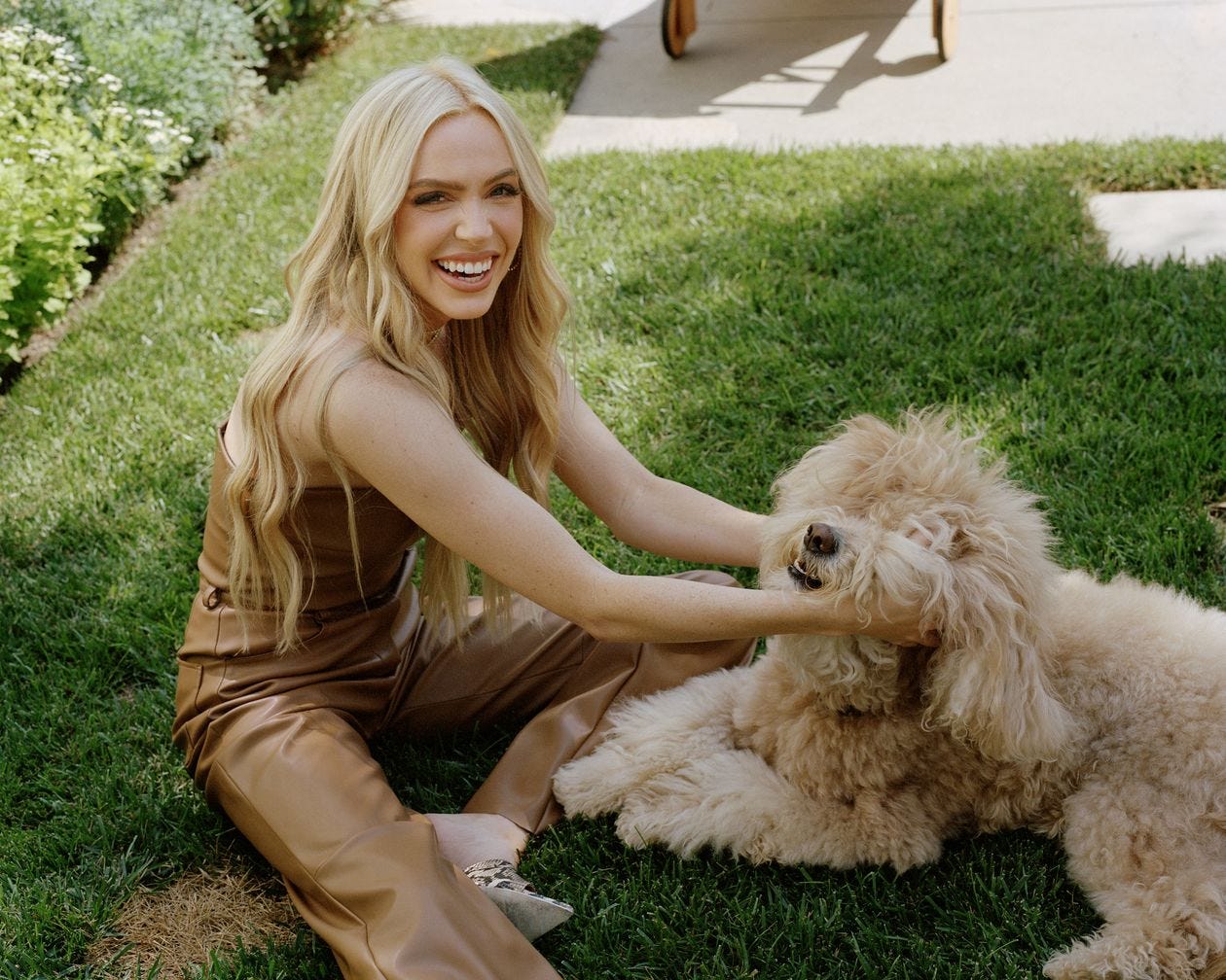 Picture of Alex Cooper, blond and smiling in brown leather pants and a leather tube top (?) sitting on a green lawn and scritching a very fluffy pup. Picture of Alex Cooper, blond and smiling in brown leather pants and a leather tube top (?) sitting on a green lawn and scritching a very fluffy pup.