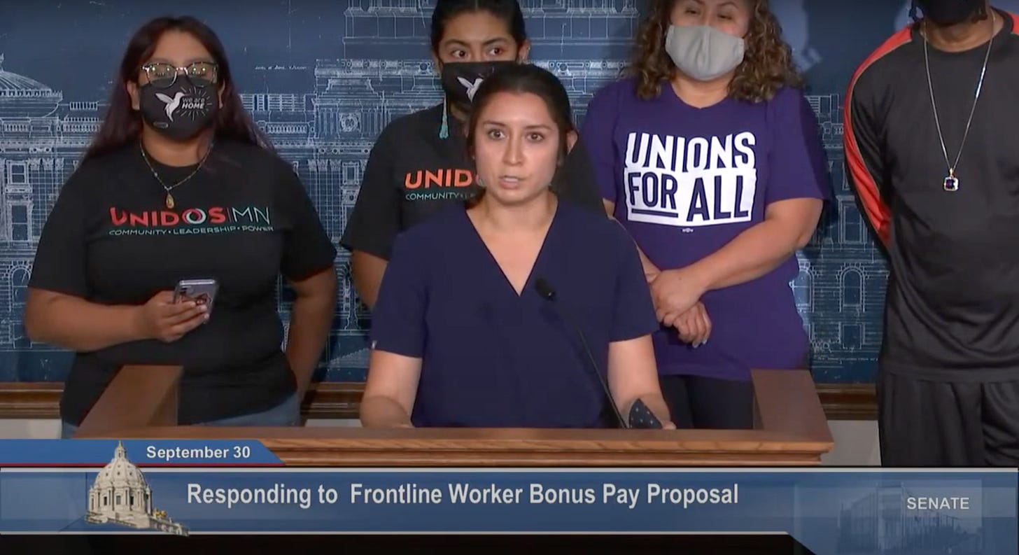 A white woman with brown hair in navy scrubs stands behind a podium, behind her people in graphic tees reading Unidos MN and Union for All