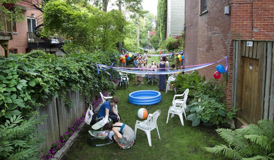 This photo shows a long narrow patch of grass between two brick apartment buildings. Adults and small children appear to be having a party, seated on white plastic chairs, with a kiddie pool on the ground, and colorful streamers and balloons tied to the building wall and the fence at the left of the frame. Leaves, vines, and trees are visible over the top of the fence. 