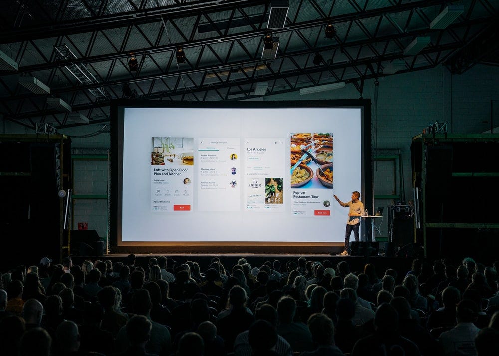 person discussing while standing in front of a large screen in front of people inside dim-lighted room person discussing while standing in front of a large screen in front of people inside dim-lighted room