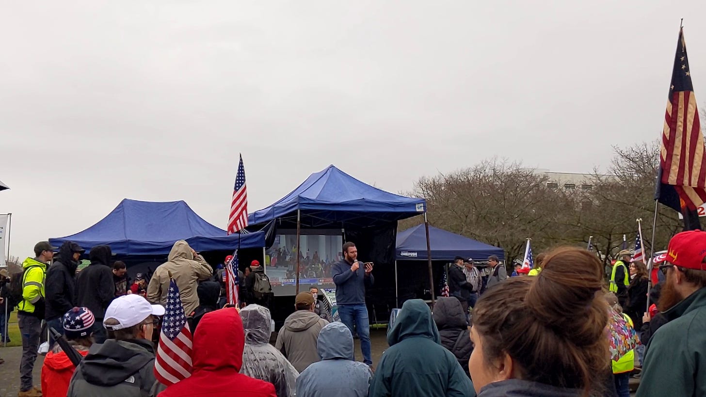 A crowd of 30-40 people in rain gear with American flags stares at a stage covered with a blue awning. A man in a blue zip-up sweater and blue jeans with a brown beard reads from his phone. Behind him, a screen shows something that looks a bit like the national capital steps