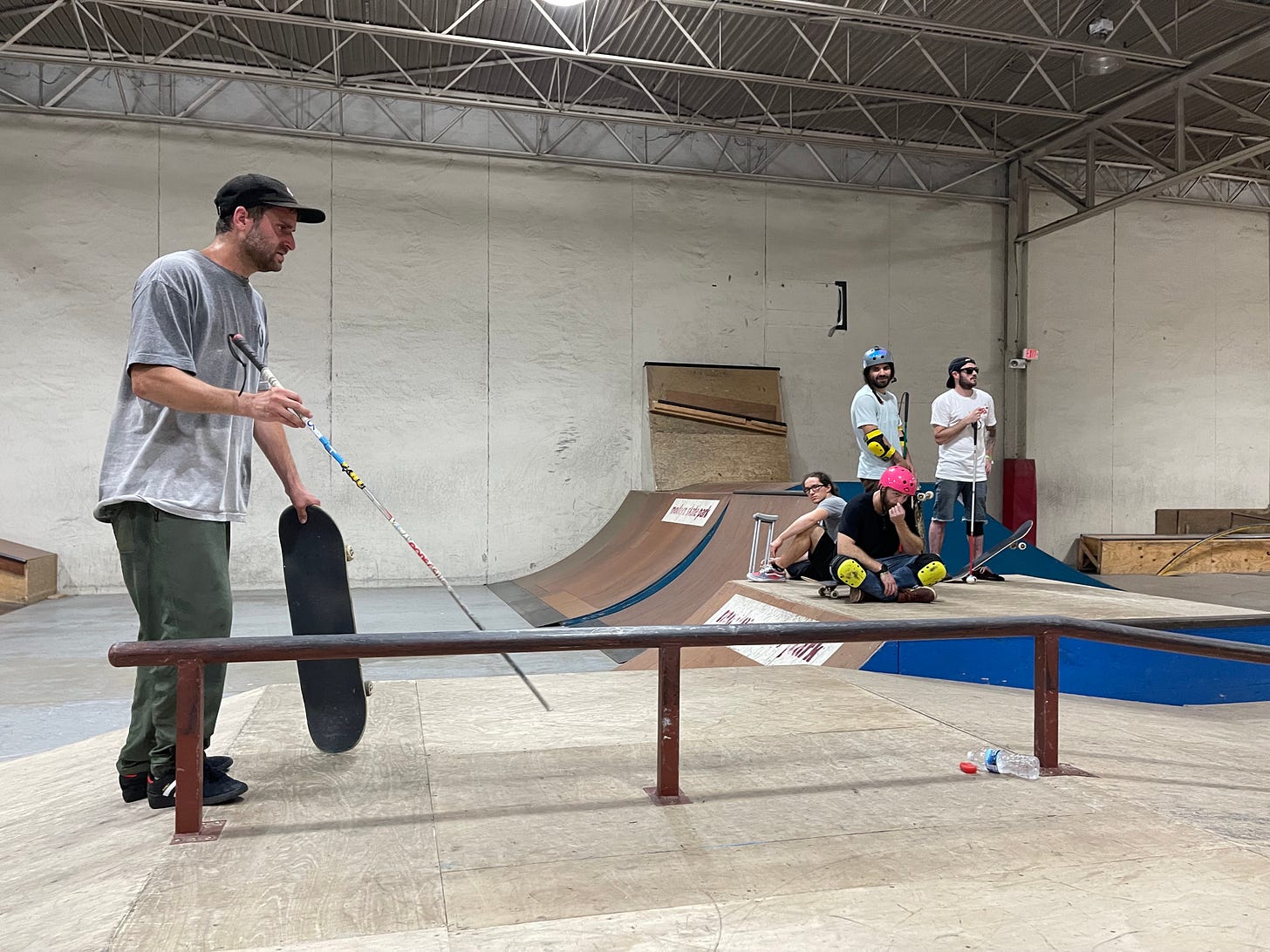 Blind skater Dan Mancina holds his skateboard as he studies the rail with his cane. 3 other blind skaters listen on in the background one of the skaters has a broken leg