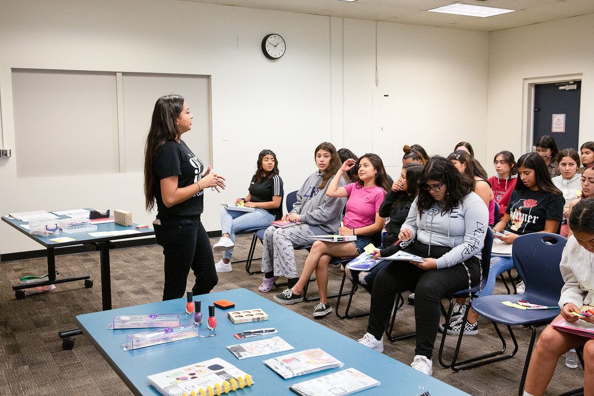 A teacher stands before a classroom full of students, with a table holding nail polish and other prizes in the background. A teacher stands before a classroom full of students, with a table holding nail polish and other prizes in the background.
