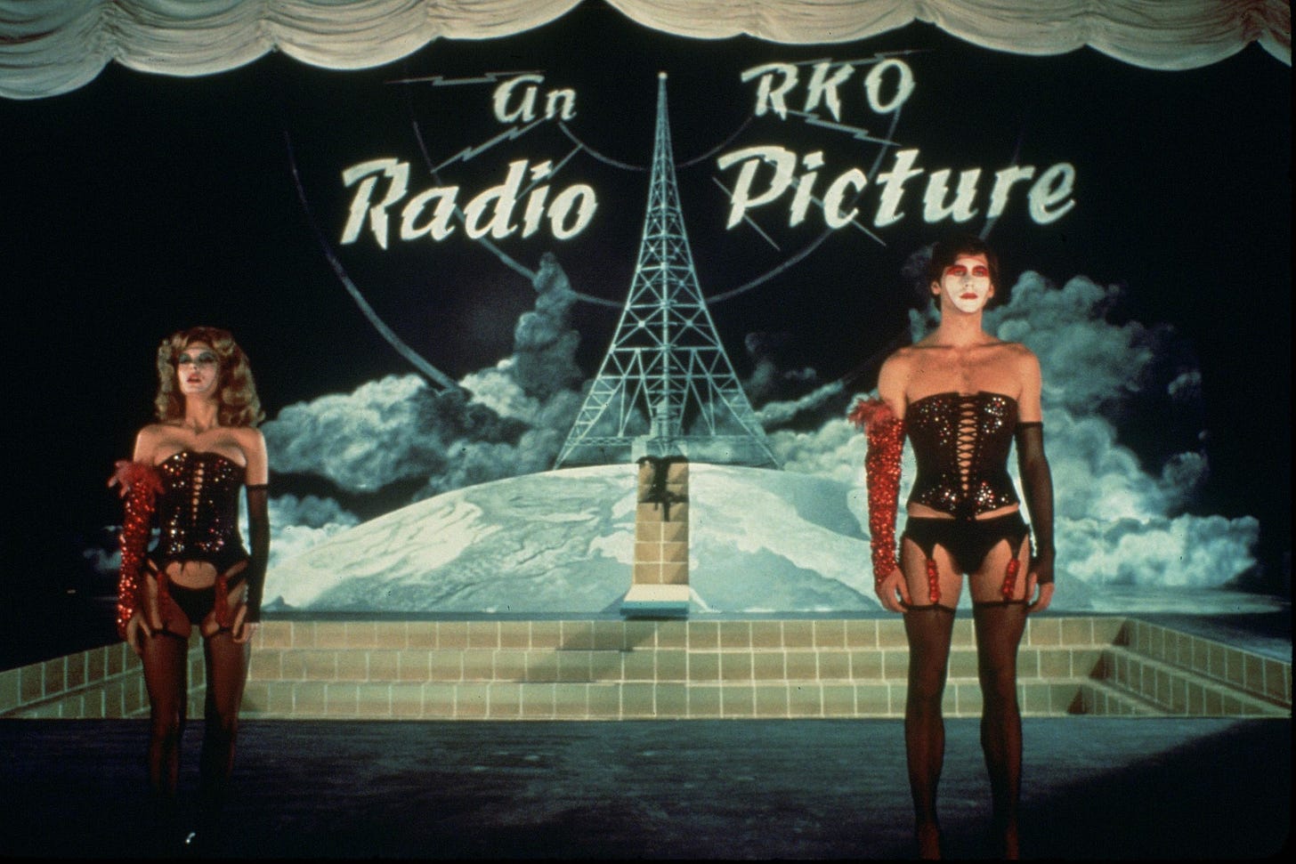 Brad and Janet stand in corsets and garters with makeup in front of an RKO Radio Picture sign