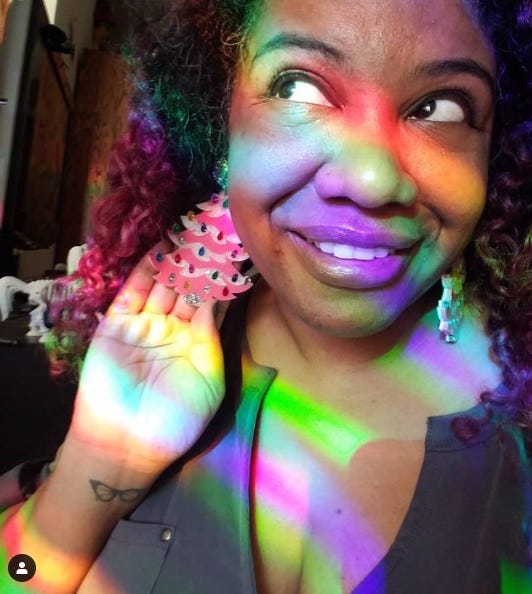 Photo of Patricia wearing large earrings that are acrylic pink Christmas trees with lots of glitter. She is covered in rainbows made by suncatchers in her window. Photo of Patricia wearing large earrings that are acrylic pink Christmas trees with lots of glitter. She is covered in rainbows made by suncatchers in her window.