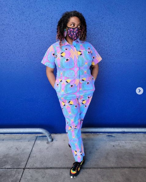Photo of Patricia standing with her hands in her pockets in front of a bright blue wall. She is wearing a multicolored jumpsuit and a tropical print face mask. She is looking to the right.