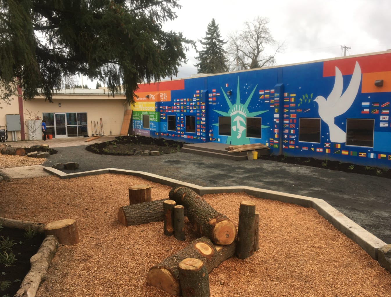 The same parking lot as above is pictured, but most of the pavement has been replaced by wood mulch, decorative tree stumps, and a colorful painting on the side of the building, featuring (from left to right) the colors of the rainbow flag, the word "peace" in many languages, flags of the world, a stylized illustration of the Statue of Liberty, and a white dove carrying an olive branch. There is fresh soil and small plants growing along the edge of the building. 