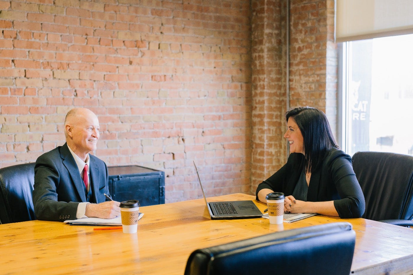 Two people are sitting at a conference table, a man and a woman. Both are dressed formally, with the man wearing a suit and tie and the woman in formal dress. The woman has a laptop in front of her while the man has a notepad. Two people are sitting at a conference table, a man and a woman. Both are dressed formally, with the man wearing a suit and tie and the woman in formal dress. The woman has a laptop in front of her while the man has a notepad.