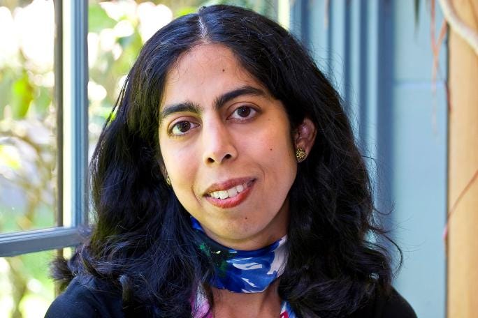 A photo of author Sheela Chari looking at the camera, wearing a navy blue sweater and a blue and purple scarf, standing in front of a clear window with light blue trim, with trees and green leaves in the background. A photo of author Sheela Chari looking at the camera, wearing a navy blue sweater and a blue and purple scarf, standing in front of a clear window with light blue trim, with trees and green leaves in the background.