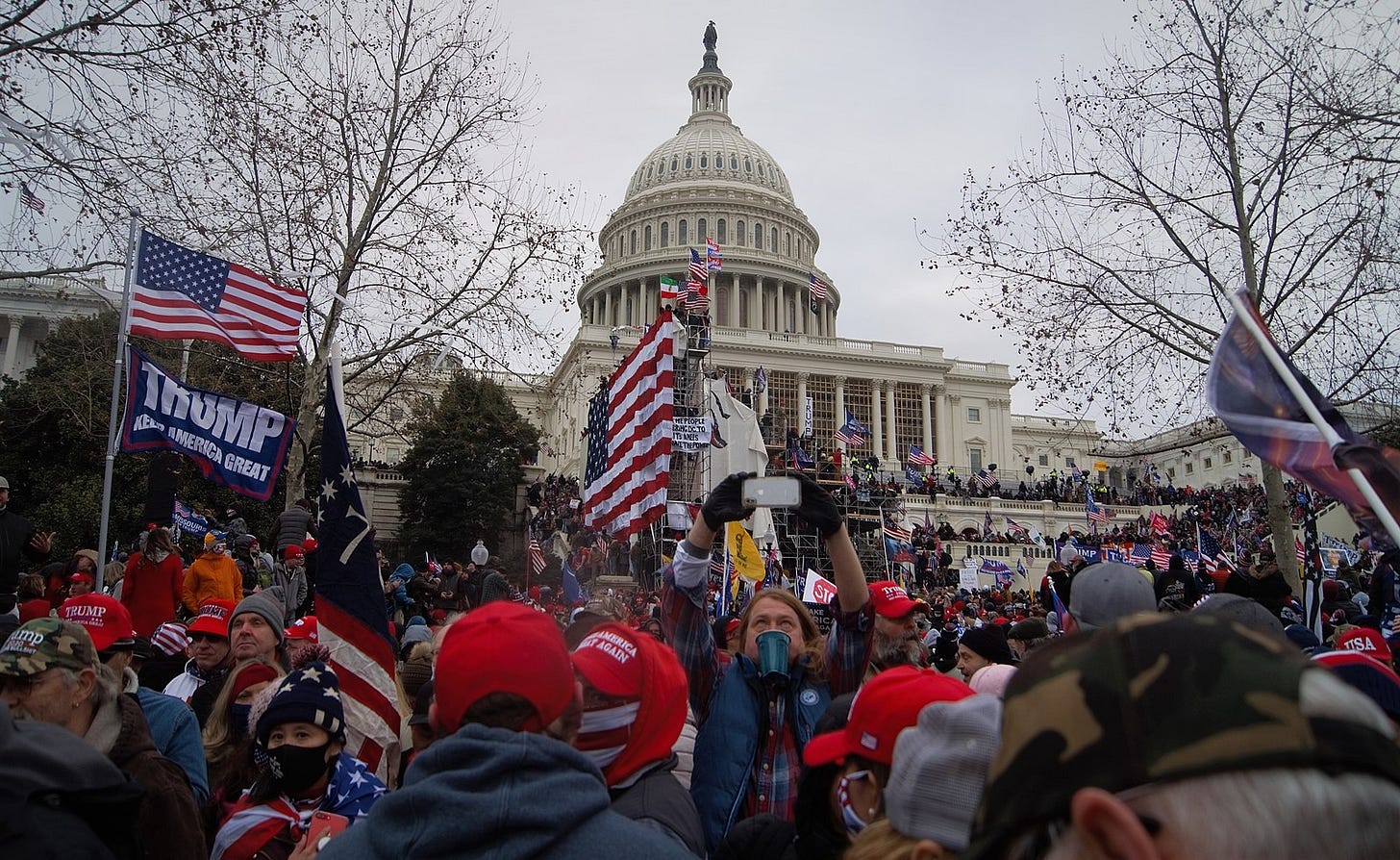 A crowd of protestors, many wearing red hats and waving American flags, is standing in front of the Capitol building. One person is also flying a Trump, Make America Great flag. A white man is holding up his phone to take a video of the scene. Protesters have climbed up the walls of the Capitol.
