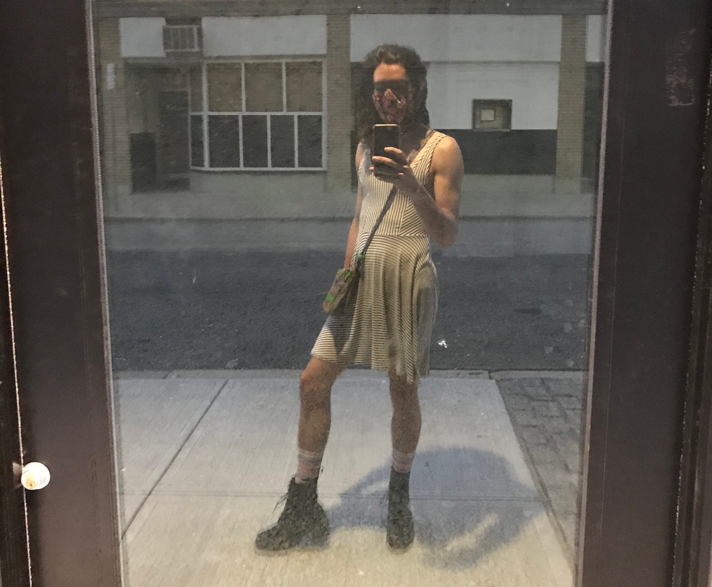 A somewhat blurry mirror-esque selfie taken in a very reflective glass door on an empty industrial-looking downtown Providence street. Nico stands in the middle, body cheated a bit to the side with one leg out, wearing a black and white striped dress that cuts off just above the knee. They have big Doc Martens on and high socks, a colorful satchel over one shoulder and a dark face mask. They raise on arm in front of them, muscles lightly flexed, to hold the phone taking the picture. Their long hair is swept over to one side, their face mostly obscured as they look down at the screen.