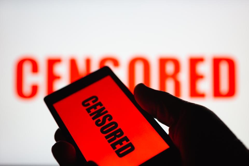 A smartphone in hand displaying the ″censored″ text. Same text in red blurred on white background. The concept of censorship on popular social networks. Restricted access to internet. Shallow DOF
