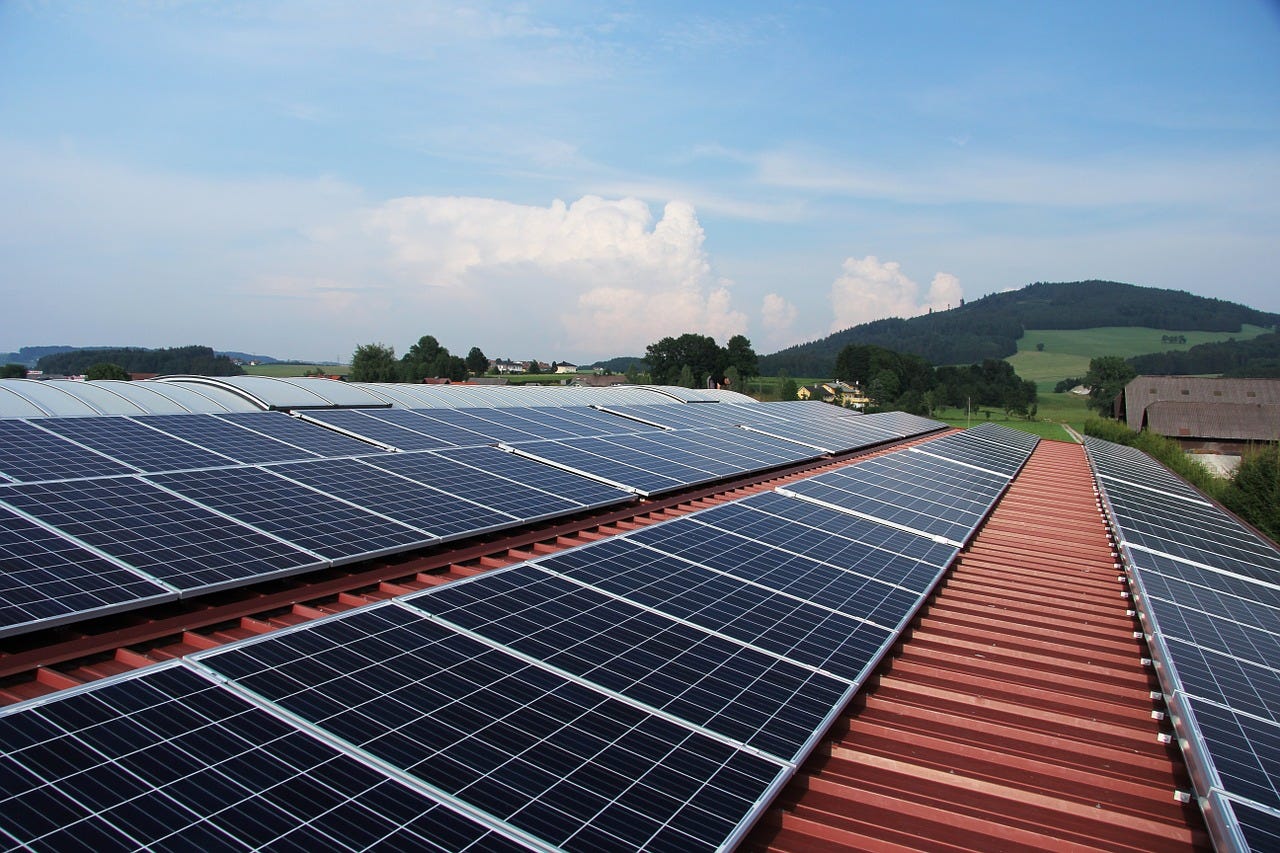 An array of solar panels on a red roof with green hills and a blue sky in the background. An array of solar panels on a red roof with green hills and a blue sky in the background.