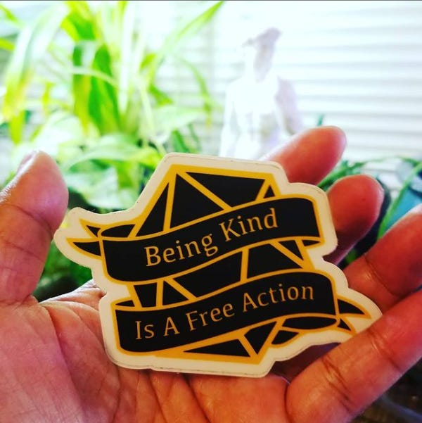 Photo of my hand holding a sticker. The sticker is in the shape of a black D20 and has the text, “Being Kind is a Free Action.” In the background is a replica of the statue of David and some plants.