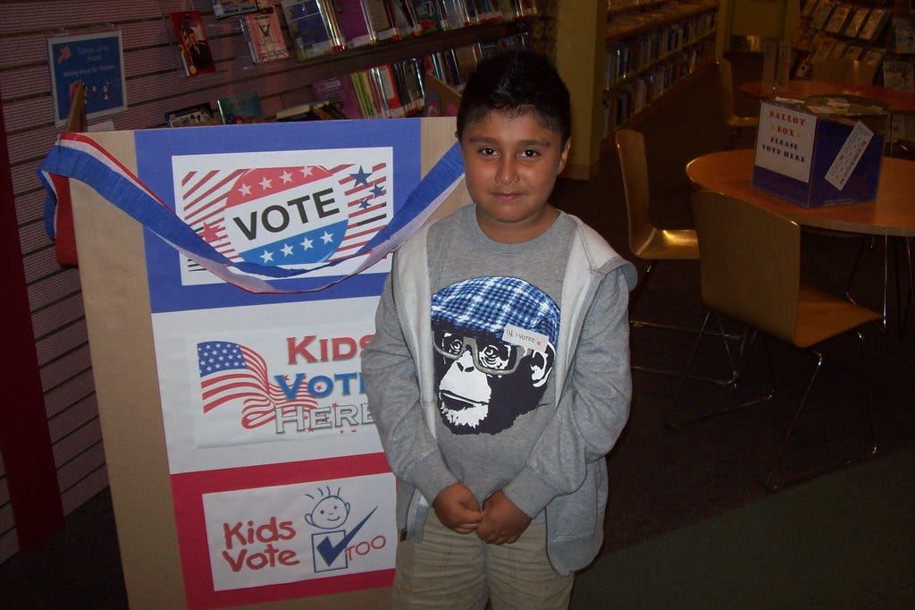 A young boy stands next to a Kids Vote display at the San Jose Pubic Library.