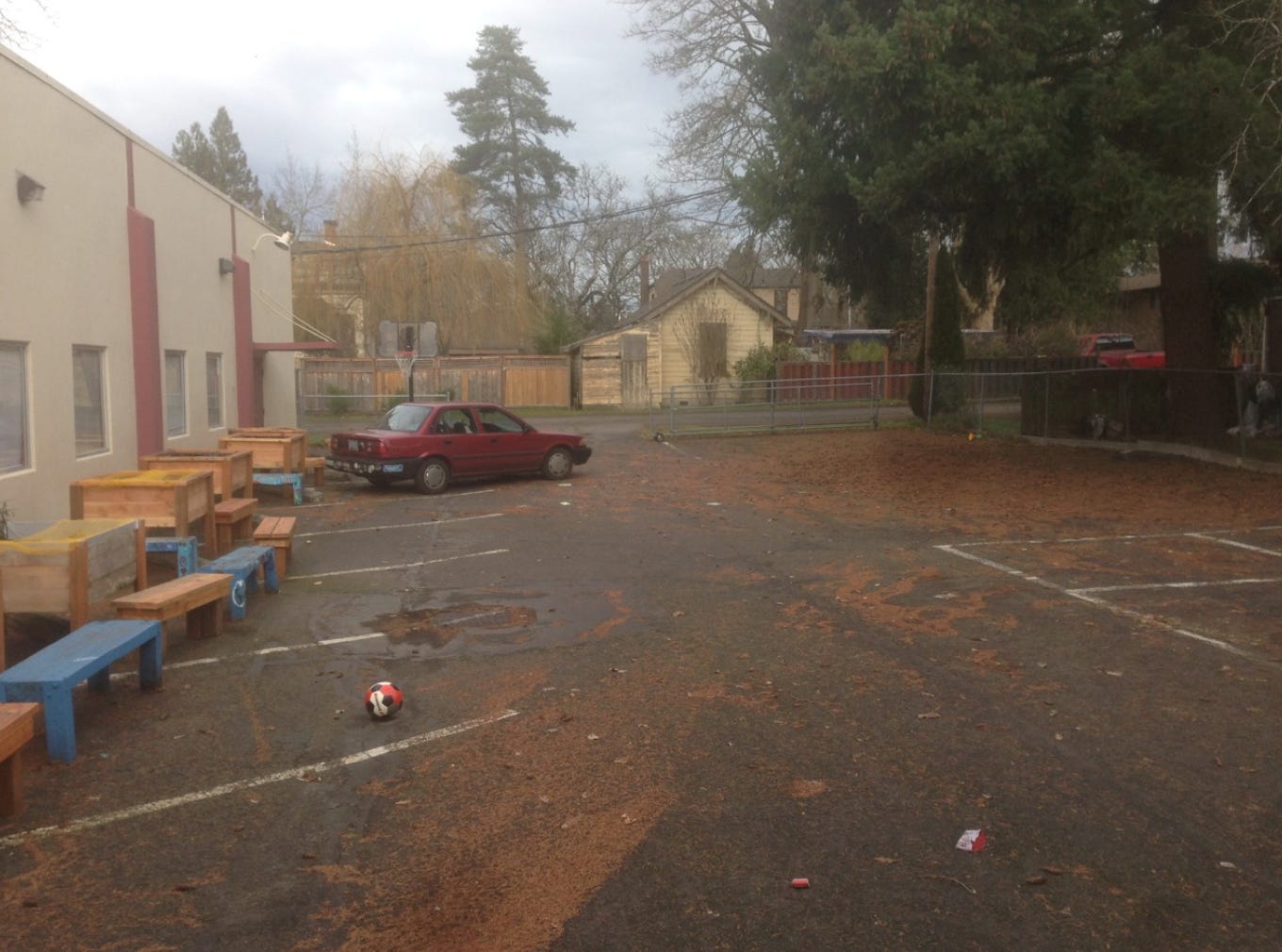 A photograph of a square parking lot, covered with asphalt. There is one small red sedan parked in the back left corner, and a row of wooden planter boxes and benches along the left site. A lone soccer ball sits in the near left corner of the frame, and there are pine needles scattered throughout the lot. The parking lot appears to open to a residential street.