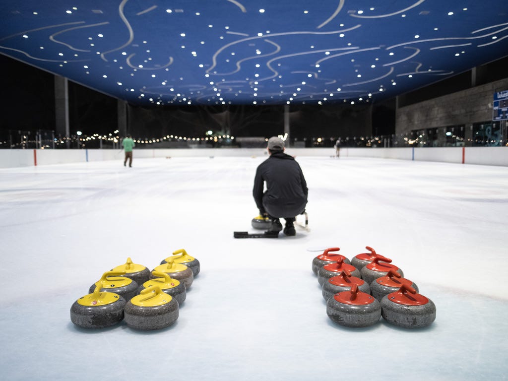 A man in a baseball cap faces away from the camera, preparing to slide a large curling stone down an ice rink. Curling stones for each team are set up behind him.