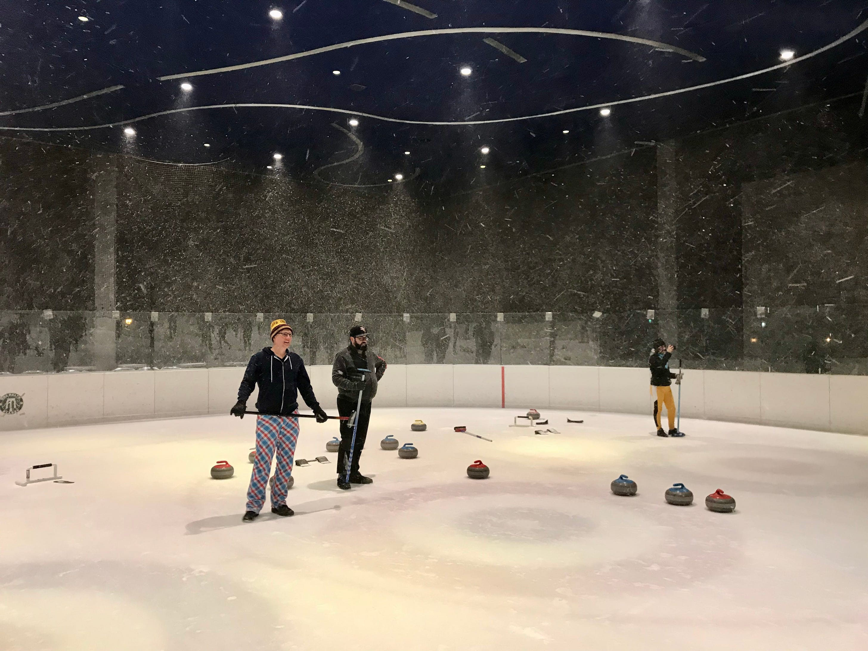 Three curlers stand on an ice rink, surrounded by curling stones, as a snowstorm rages around them.