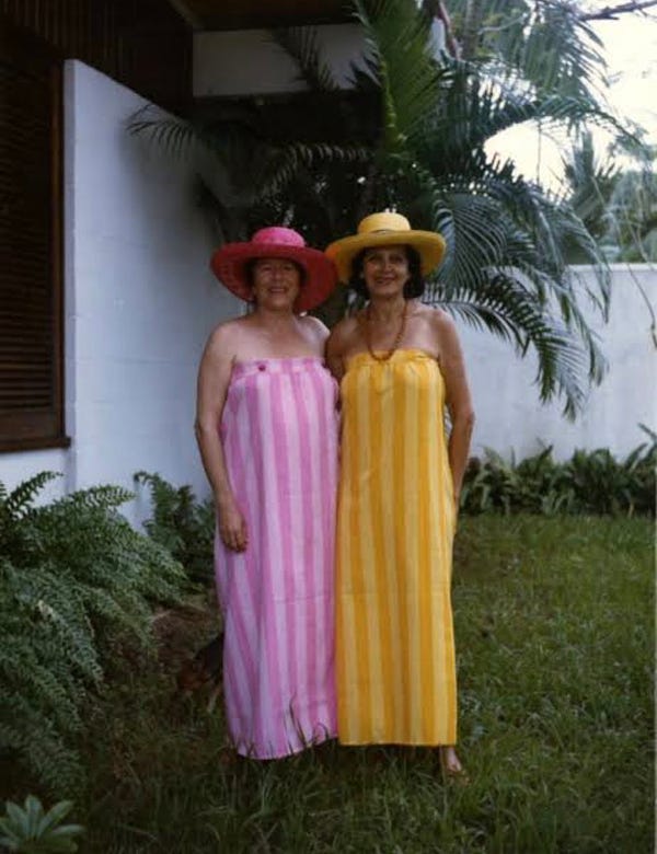 Fotografia. Em um jardim gramado, duas mulheres em pé, uma ao lado da outra. À esquerda, a escritora Hilda Hilst, vestindo um vestido tomara que caia listrado em tons de rosa e um chapéu rosa. À direita, a escritora Lygia Fagundes Telles, vestindo um vestido igual, em tons de amarelo, chapéu amarelo e um colar cor de âmbar. Ao fundo, um muro branco.