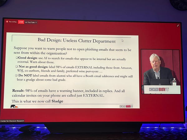 A picture of Richard Thaler presenting a slide on bad design, specifically useless clutter. His example is warnings about phishing emails, such as flagging anything from any external sender. If 98% of emails have a warning banner, this becomes “sludge”.