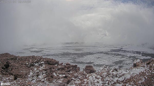 Snow covers a brown volcanic rock landscape and low clouds touch the ground
