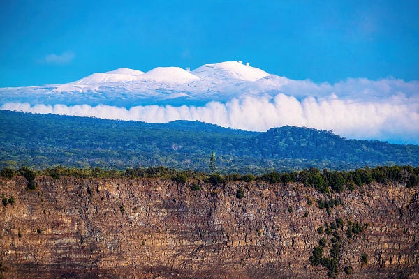 3 Hawaiian volcanoes piled on top of each other like layer cake: snowy Mauna Kea on top, a forested slope of Mauna Loa and the brown crater walls of Kilauea all in frame in one spectacular photo. 